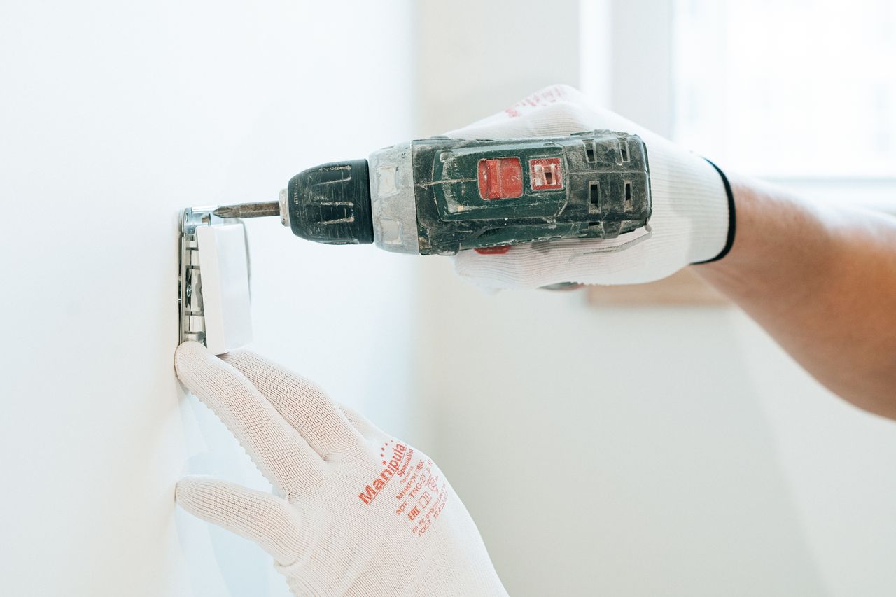 Gloved hands using a drill to install a power outlet on a wall, focusing on precision and safety.